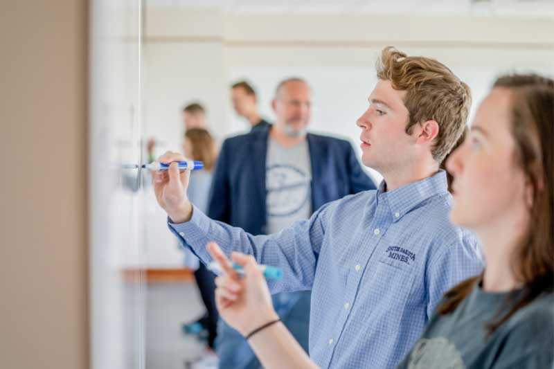 Students collaborating and writing on a whiteboard in a classroom.