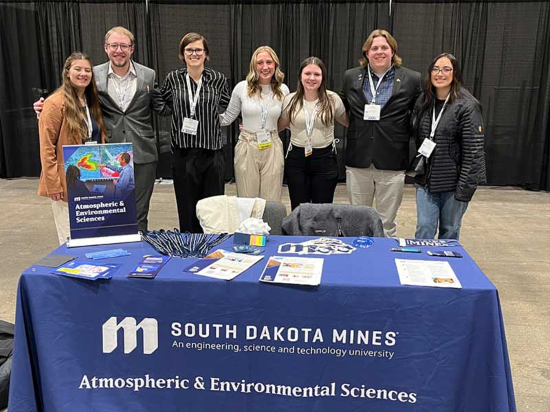 Group of students standing behind a South Dakota Mines Atmospheric and Enviromental Sciences display table.